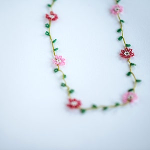 Pu&ograve; includere: Una delicata collana di perline con fiori rosa e rossi e foglie verdi. La collana &egrave; realizzata con piccole perline ed &egrave; infilata su un filo sottile.