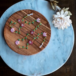 Pu&ograve; includere: Una collana di perline con fiori rosa e verdi su un piatto di legno. La collana &egrave; su una superficie di marmo.