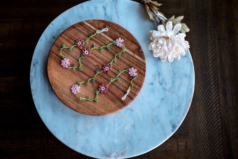 Pu&ograve; includere: Una collana di perline con fiori rosa e verdi su un piatto di legno. La collana &egrave; su una superficie di marmo.