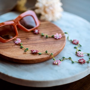 Pu&ograve; includere: Un paio di occhiali da sole arancioni con una catena per occhiali a perline floreali. La catena presenta perline rosa e verdi a forma di fiori e foglie.