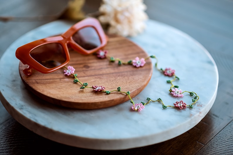 Pu&ograve; includere: Un paio di occhiali da sole arancioni con una catena per occhiali a perline floreali. La catena presenta perline rosa e verdi a forma di fiori e foglie.