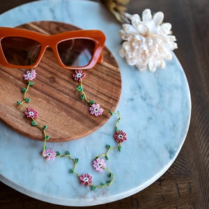 Pu&ograve; includere: Occhiali da sole arancioni con una catena floreale di perline. La catena &egrave; realizzata con perline rosa e verdi e presenta piccoli disegni di fiori. Gli occhiali da sole e la catena sono su una superficie di legno e marmo.