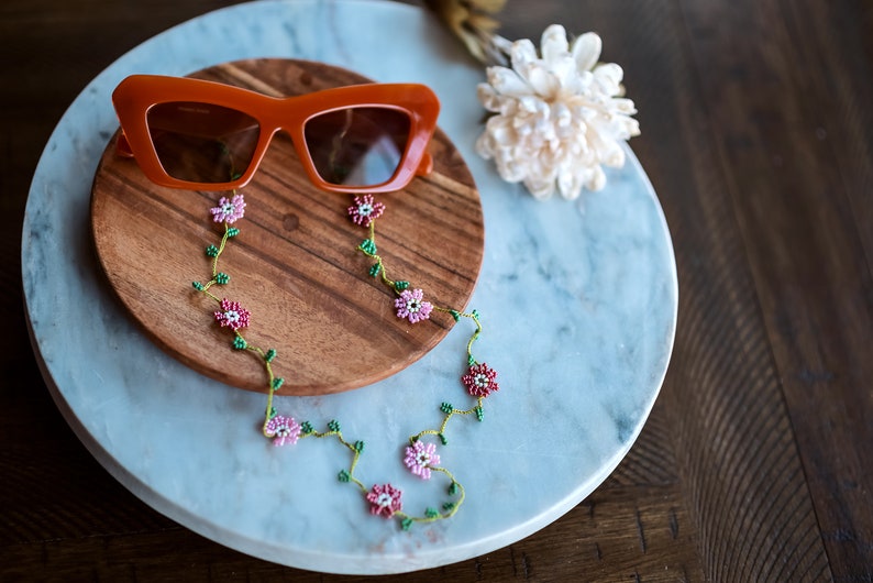 Pu&ograve; includere: Occhiali da sole arancioni con una catena floreale di perline. La catena &egrave; realizzata con perline rosa e verdi e presenta piccoli disegni di fiori. Gli occhiali da sole e la catena sono su una superficie di legno e marmo.