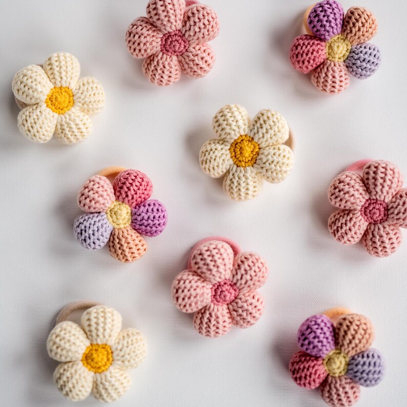 K&ouml;nnte beinhalten: Ein Set mit zehn bunten geh&auml;kelten Blumenhaargummis. Die Haargummis sind in verschiedenen Farben gehalten, darunter Rosa, Gelb und Lila. Jedes Haargummi hat ein Blumendesign mit einem Knopf in der Mitte.