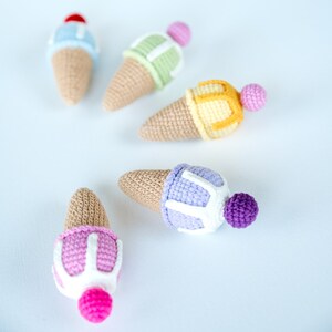 May include: Five crocheted ice cream cones in different colors. The cones are made of beige yarn and the ice cream is made of different colored yarns. The ice cream cones are arranged in a semi-circle on a white background.
