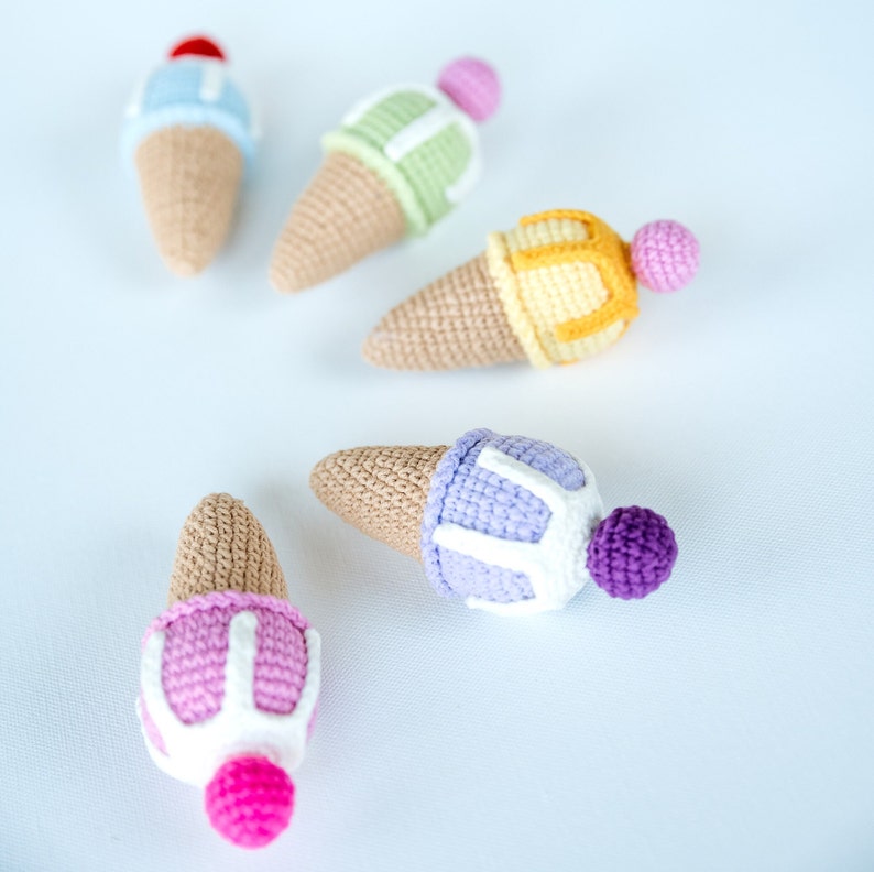 May include: Five crocheted ice cream cones in different colors. The cones are made of beige yarn and the ice cream is made of different colored yarns. The ice cream cones are arranged in a semi-circle on a white background.