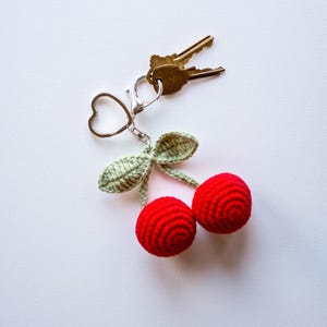 May include: A handmade cherry keychain with two red crocheted cherries, green leaves, and a silver-tone heart-shaped keyring. The keychain is attached to two gold keys, set against a white background.