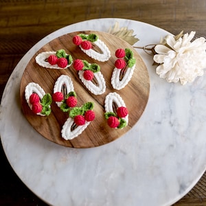 Puede incluir: Ocho pinzas para el pelo de ganchillo con detalles rojos y verdes, que se asemejan a fresas, dispuestas sobre una superficie de madera.