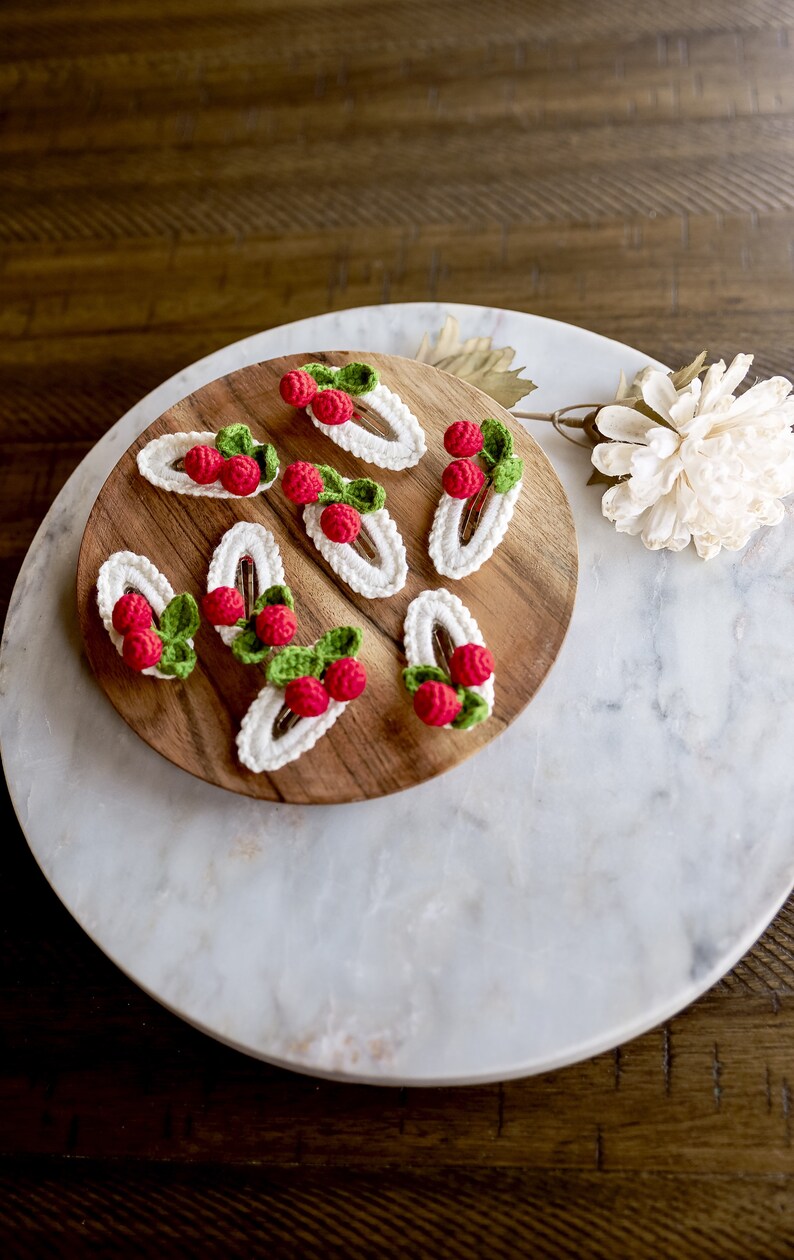 Puede incluir: Ocho pinzas para el pelo de ganchillo con detalles rojos y verdes, que se asemejan a fresas, dispuestas sobre una superficie de madera.