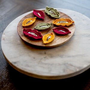 May include: A set of eight crocheted leaf-shaped hair clips in red, yellow, and green. The clips are arranged on a wooden plate, which is on a marble surface.