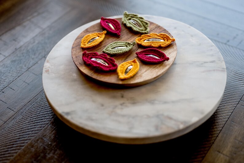 May include: A set of eight crocheted leaf-shaped hair clips in red, yellow, and green. The clips are arranged on a wooden plate, which is on a marble surface.