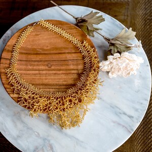 Puede incluir: Un collar de cuentas doradas con un diseño delicado e intrincado. El collar se muestra en un plato de madera, que está sobre una superficie de mármol blanco.
