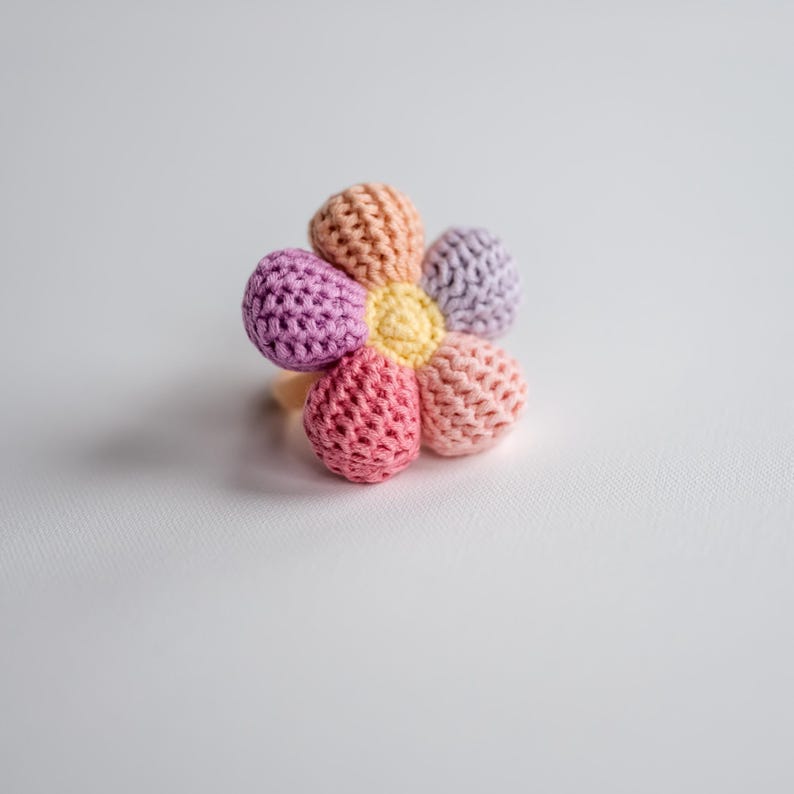 K&ouml;nnte beinhalten: Ein geh&auml;keltes Spielzeug in Form einer Blume mit rosa, lila und gelben Bl&uuml;tenbl&auml;ttern. Das Spielzeug ist aus weichem Garn gefertigt und perfekt f&uuml;r Babys und Kleinkinder.