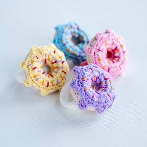 May include: Four crocheted donuts in different colors: yellow, blue, pink, and purple. Each donut has a white hole in the center and colorful sprinkles.