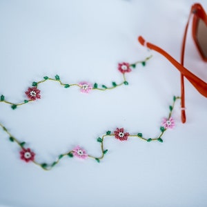 Pu&ograve; includere: Una delicata collana di perline con fiori rosa e bianchi e foglie verdi. La collana &egrave; adagiata su una superficie bianca accanto a un paio di occhiali da sole arancioni.