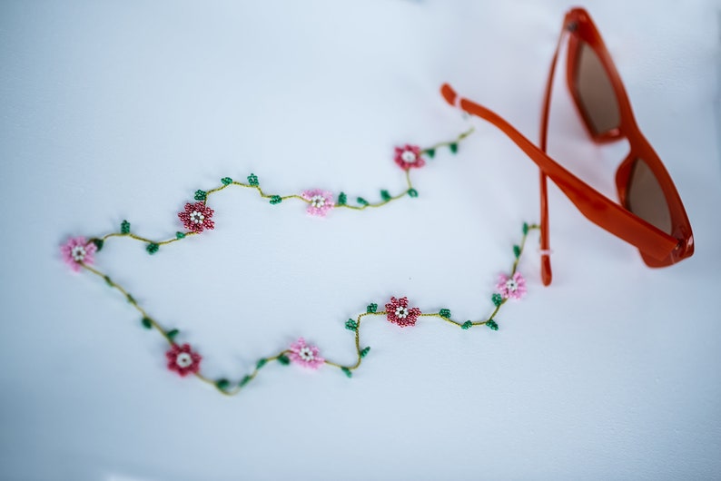 Pu&ograve; includere: Una delicata collana di perline con fiori rosa e bianchi e foglie verdi. La collana &egrave; adagiata su una superficie bianca accanto a un paio di occhiali da sole arancioni.