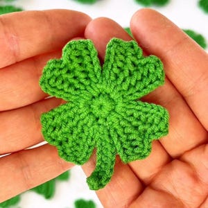 Puede incluir: Un trébol de cuatro hojas de ganchillo hecho a mano, de color verde brillante. El trébol se sostiene en manos abiertas, mostrando las puntadas detalladas y la textura del hilo. El trébol es un símbolo de buena suerte y es perfecto para el Día de San Patricio.