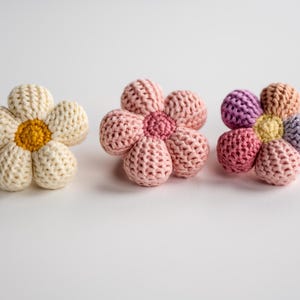 K&ouml;nnte beinhalten: Drei geh&auml;kelte Blumenspielzeuge. Eines ist wei&szlig; mit gelber Mitte, eines ist rosa mit rosa Mitte und eines ist mehrfarbig mit gelber Mitte.