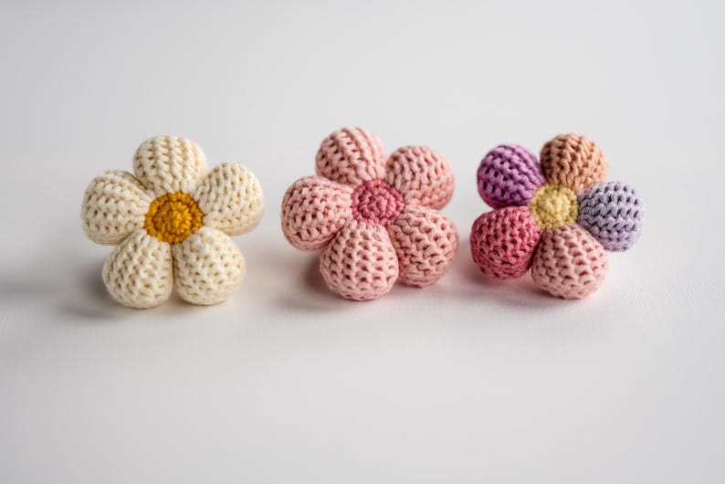 K&ouml;nnte beinhalten: Drei geh&auml;kelte Blumenspielzeuge. Eines ist wei&szlig; mit gelber Mitte, eines ist rosa mit rosa Mitte und eines ist mehrfarbig mit gelber Mitte.