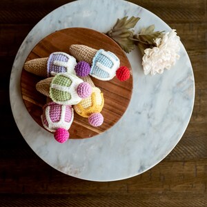 May include: A group of six crocheted ice cream cones in various colors, including pink, purple, yellow, green, and blue, are arranged on a wooden cutting board. The cones are topped with colorful crocheted balls.