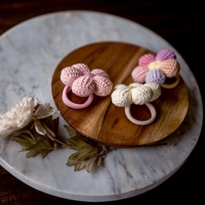 K&ouml;nnte beinhalten: Drei geh&auml;kelte Blumenhaargummis in Rosa, Wei&szlig; und Lila auf einer Holzoberfl&auml;che. Die Haargummis befinden sich auf einer Marmorfl&auml;che.