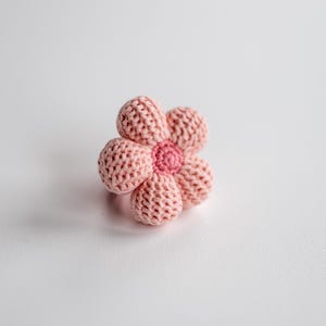 K&ouml;nnte beinhalten: Eine rosa geh&auml;kelte Blume mit einer rosa Mitte. Die Blume liegt auf einer wei&szlig;en Oberfl&auml;che.
