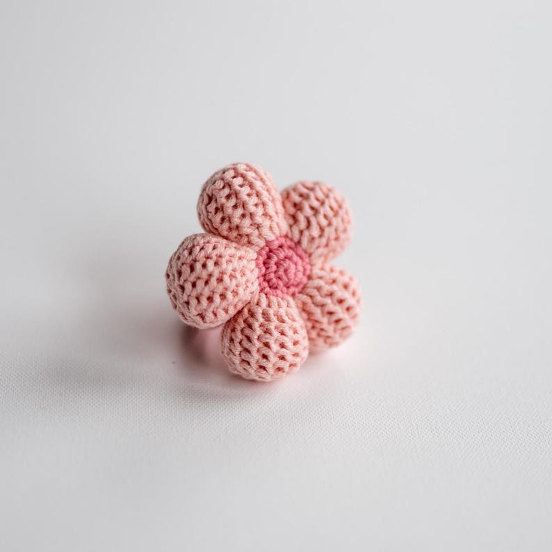 K&ouml;nnte beinhalten: Eine rosa geh&auml;kelte Blume mit einer rosa Mitte. Die Blume liegt auf einer wei&szlig;en Oberfl&auml;che.