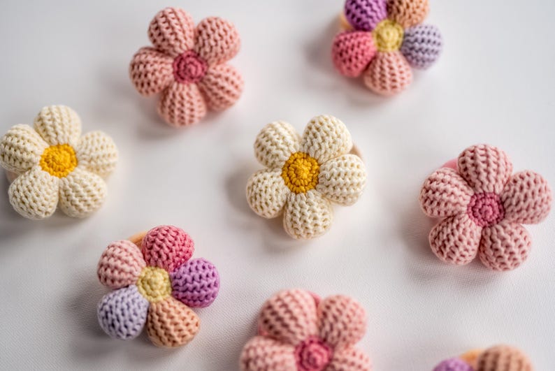 K&ouml;nnte beinhalten: Ein Set mit sechs geh&auml;kelten Blumenhaargummis in verschiedenen Farben, darunter Rosa, Wei&szlig; und Lila. Die Blumen haben ein schlichtes, g&auml;nsebl&uuml;mchenartiges Design mit einem Knopf in der Mitte.