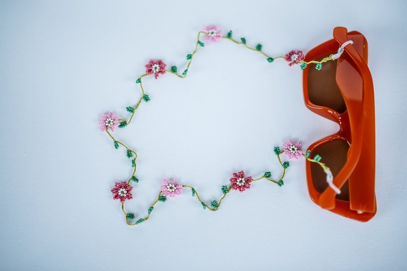Pu&ograve; includere: Un paio di occhiali da sole arancioni con una catena di fiori perlati. La catena ha fiori rosa e bianchi con foglie verdi.
