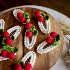 Puede incluir: Seis pinzas para el pelo de crochet blanco con frambuesas de crochet rojo y verde. Las pinzas est&aacute;n dispuestas sobre una superficie de madera.