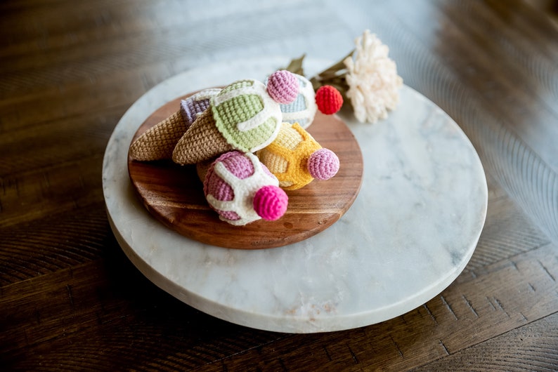 May include: A set of five crocheted ice cream cones in various colors, including pink, yellow, green, and white, displayed on a wooden platter atop a marble surface.