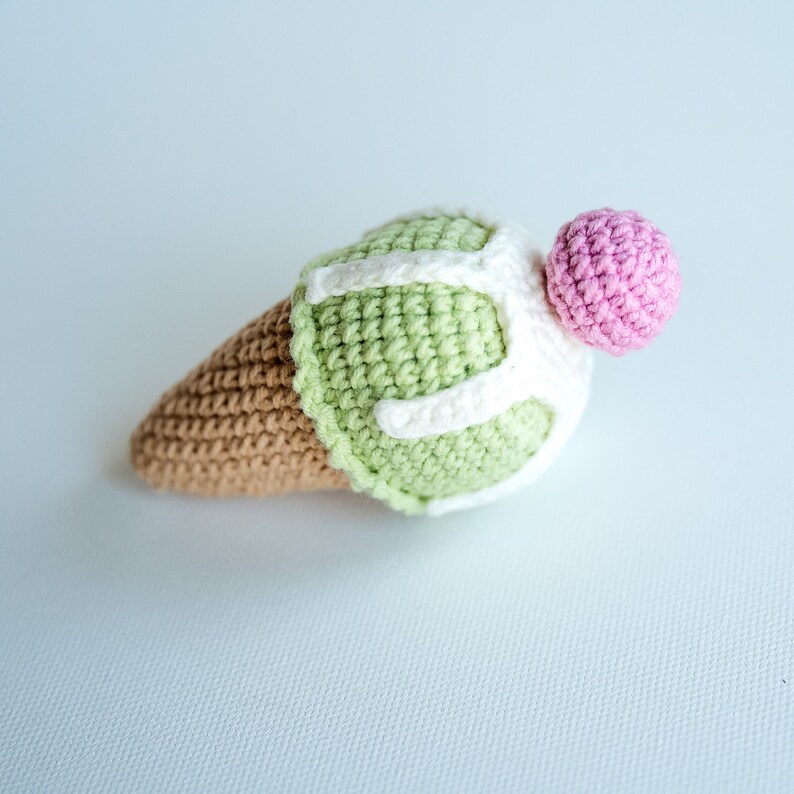 May include: A crocheted ice cream cone toy with green ice cream, white frosting, and a pink cherry on top.