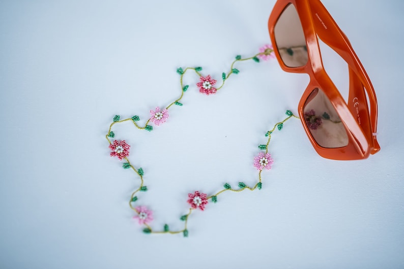 Pu&ograve; includere: Una catena di perline con fiori rosa e bianchi e foglie verdi, che forma un cerchio attorno a un paio di occhiali da sole arancioni.