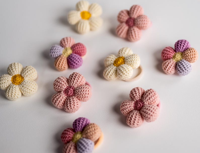 K&ouml;nnte beinhalten: Ein Set mit zehn geh&auml;kelten Blumenhaargummis in verschiedenen Rosat&ouml;nen, Wei&szlig; und Gelb. Die Haargummis sind aus weichem, strukturiertem Garn gefertigt und haben ein schlichtes Blumendesign.