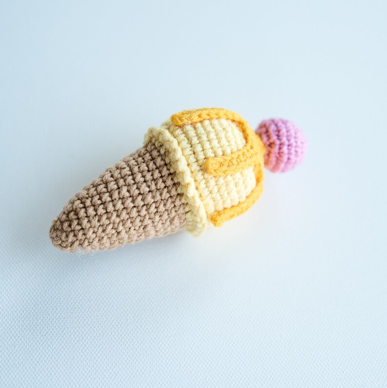 May include: A crocheted ice cream cone toy with brown, yellow, and pink yarn. The cone is brown, the ice cream is yellow with a pink cherry on top.