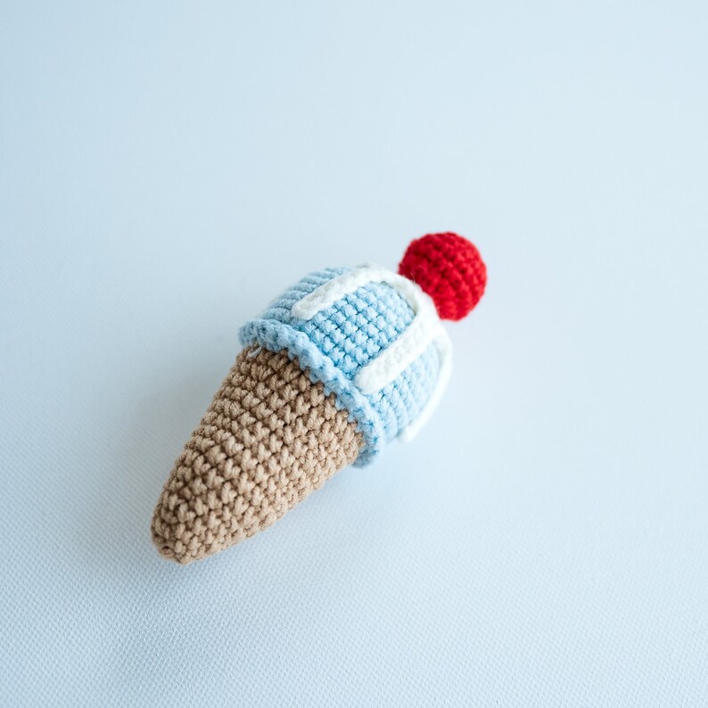 May include: A crocheted ice cream cone toy with a blue and white ice cream scoop and a red cherry on top.