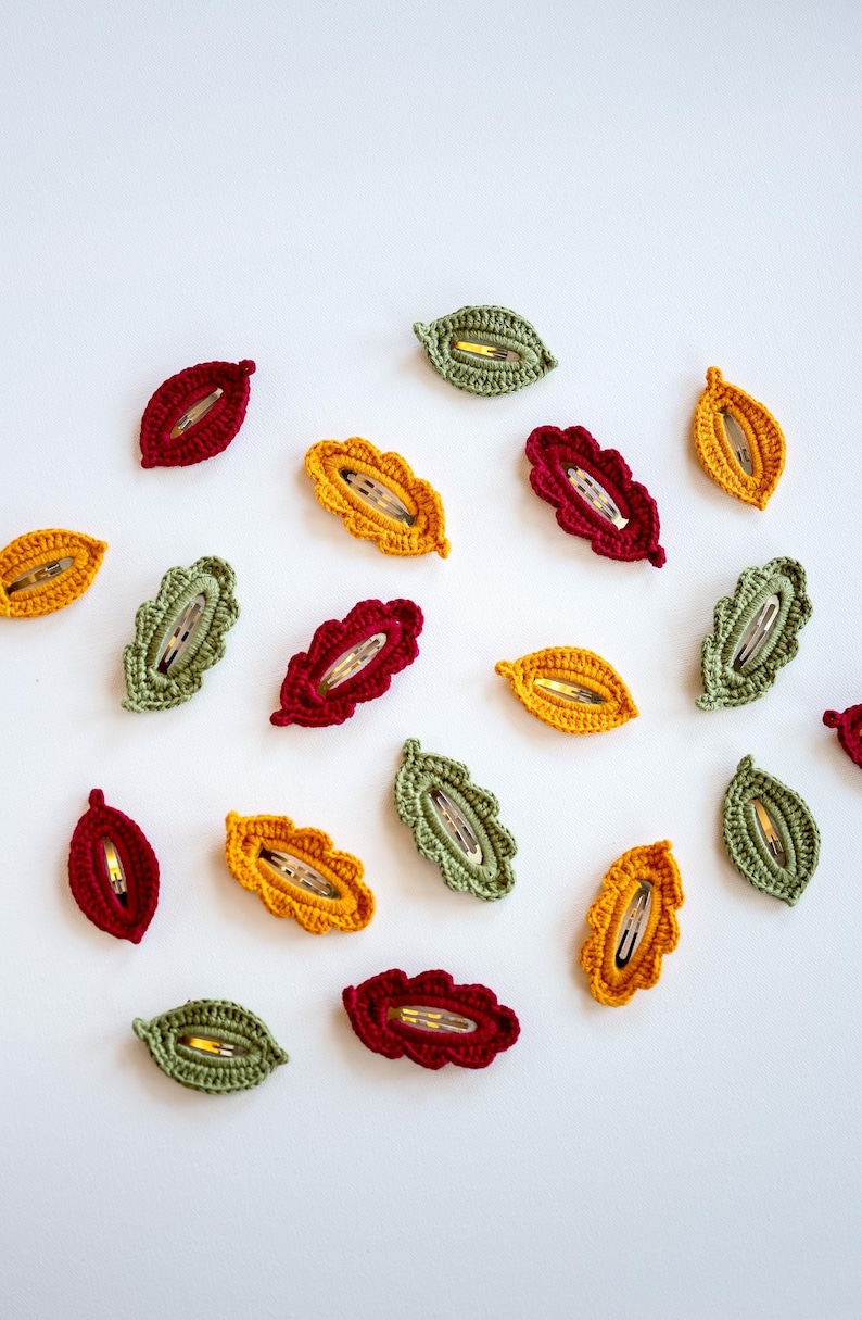 May include: A collection of crocheted leaf-shaped hair clips in shades of green, yellow, and red. The clips are arranged in a circle on a white background.