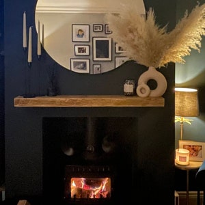 May include: A wooden mantel shelf with a white ceramic vase with a round opening, a candle, and a round mirror above a black fireplace with a fire burning inside.