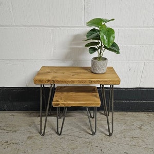 May include: A set of two wooden nesting tables with hairpin legs. The top table is larger than the bottom table. Both tables are made of reclaimed wood and have a natural finish. The tables are sitting on a concrete floor. A small potted plant is sitting on the top table.