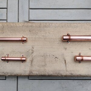 May include: Four copper drawer pulls with a brushed finish and round ends. The pulls are mounted on a wooden surface.