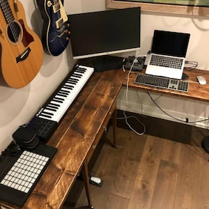 K&ouml;nnte beinhalten: Ein Ecktisch aus Holz mit einer schwarzen Tastatur, einem Laptop, einer wei&szlig;en Maus und einer schwarzen MIDI-Tastatur. Zwei Akustikgitarren h&auml;ngen an der Wand hinter dem Tisch.