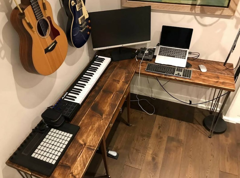 K&ouml;nnte beinhalten: Ein Ecktisch aus Holz mit einer schwarzen Tastatur, einem Laptop, einer wei&szlig;en Maus und einer schwarzen MIDI-Tastatur. Zwei Akustikgitarren h&auml;ngen an der Wand hinter dem Tisch.