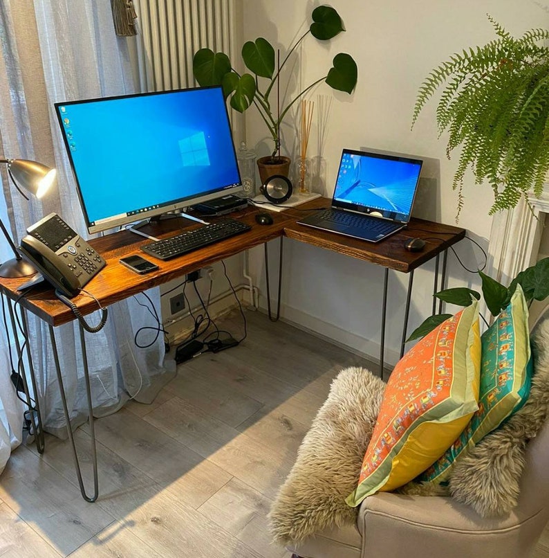 K&ouml;nnte beinhalten: Ein Ecktisch mit einer Holzplatte und schwarzen Metallbeinen. Zwei Laptops stehen auf dem Tisch, einer mit einem blauen Bildschirm und der andere mit einem schwarzen Bildschirm. Ein Telefon und eine Tastatur befinden sich ebenfalls auf dem Tisch. Im Hintergrund befindet sich eine Pflanze.
