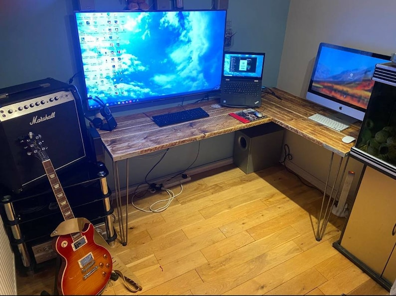 K&ouml;nnte beinhalten: Ein Ecktisch mit einer Holzplatte und Metall-Haarnadelbeinen. Auf dem Tisch stehen ein Computermonitor, ein Laptop und eine Gitarre. Im Hintergrund befinden sich au&szlig;erdem ein Lautsprecher und ein Aquarium.