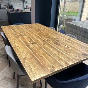 May include: A rustic wooden dining table with a natural wood finish and black metal legs. The table is set in a dining room with chairs and a view of a garden.