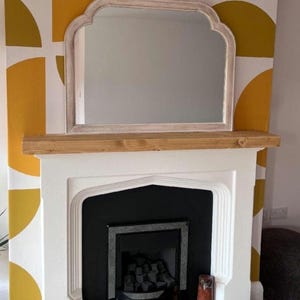 May include: A wall-mounted mirror featuring a light-coloured, weathered wood frame with a curved, arched top design. The mirror is positioned directly above a thick, natural-toned wooden mantelpiece that sits atop a white fireplace surround.