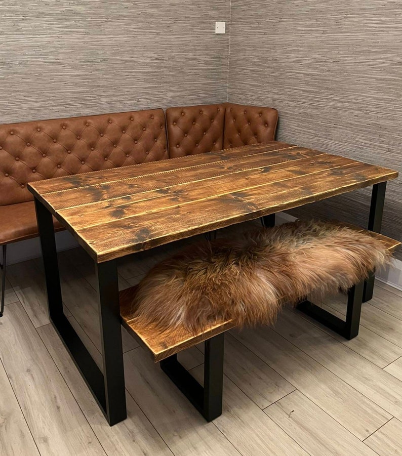 May include: A rustic wooden dining table with black metal legs and a matching bench. The table top is made of reclaimed wood with a distressed finish. The bench has a sheepskin cushion.