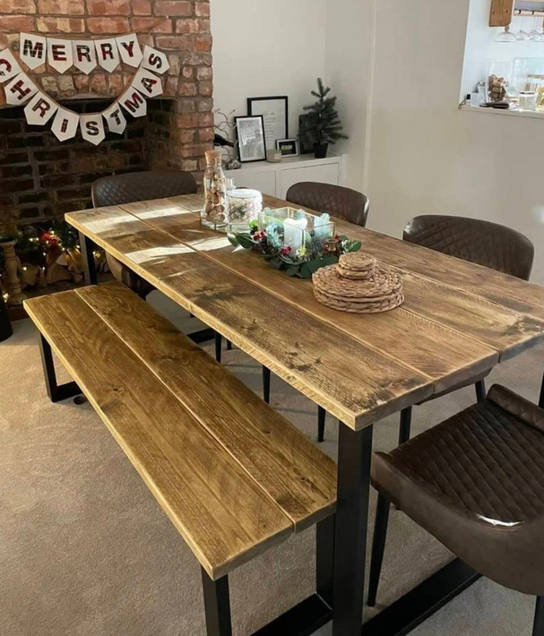 May include: A rustic wooden dining table with black metal legs and a matching bench. The table is set with a centrepiece of greenery and candles. The table is surrounded by chairs with brown leather upholstery.