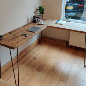 K&ouml;nnte beinhalten: Ein Ecktisch aus recyceltem Holz mit nat&uuml;rlicher Oberfl&auml;che. Der Tisch hat ein schlichtes Design mit Haarnadelbeinen und einem kleinen Regal auf der rechten Seite. Der Tisch ist f&uuml;r ein Heimb&uuml;ro mit Laptop, Telefon und Kaffeetasse auf der Oberfl&auml;che eingerichtet.