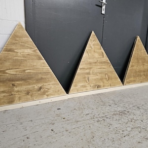 May include: Three wooden triangle shelves in various sizes, stained in a light brown colour, arranged in a row on a grey concrete floor.